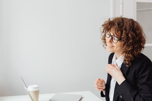 Caucasian woman with curly hair and glasses smiling while working at a desk in an office setting.
